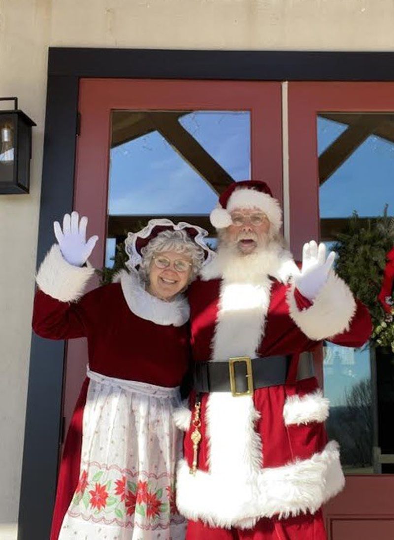 Santa John and Mrs. Claus waving from the front door of a log cabin