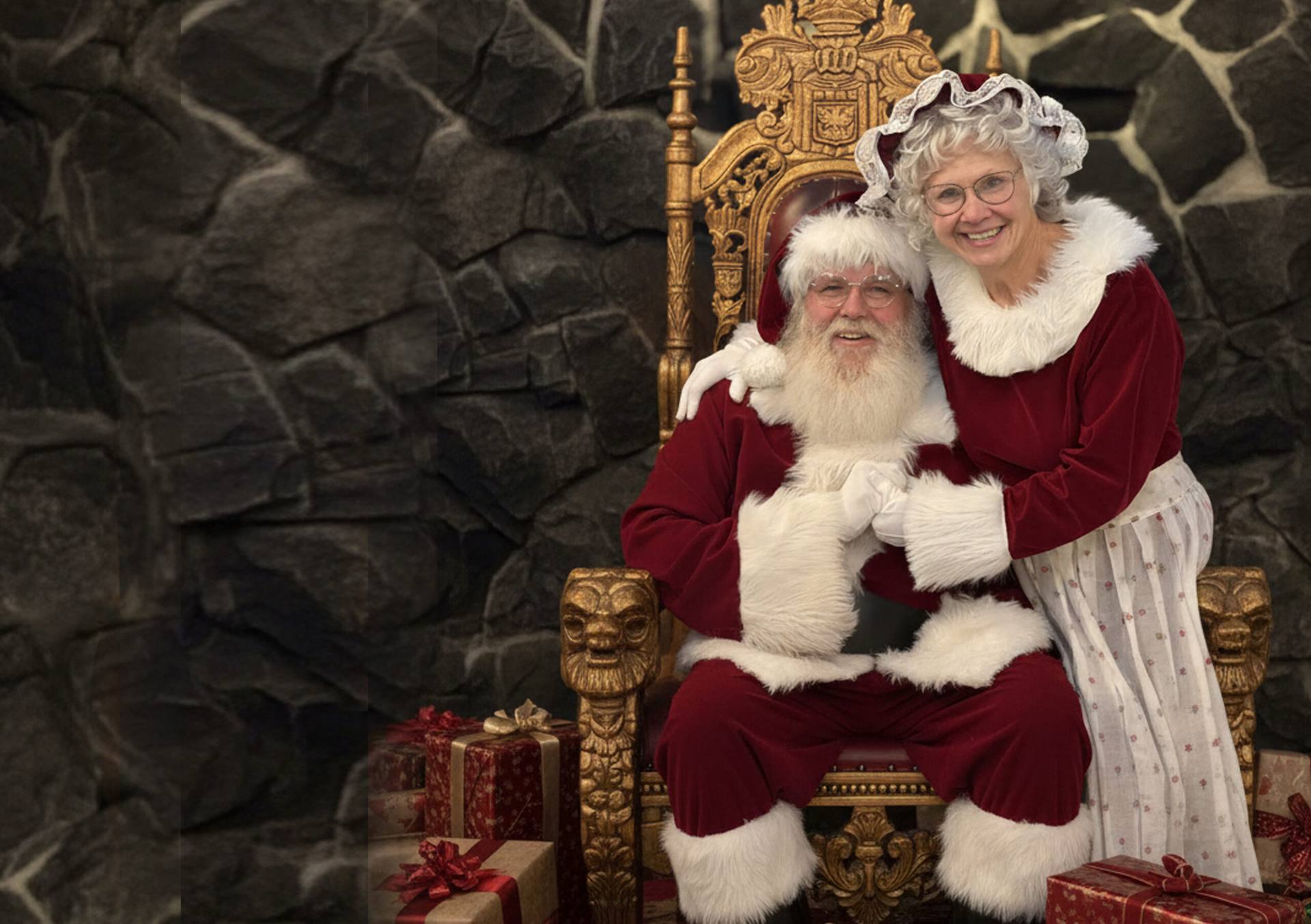 Santa John and Mrs. Claus seated together on a golden throne beside wrapped presents